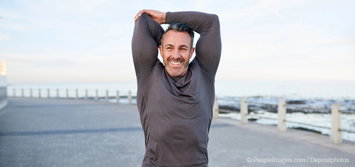 Reifer Mann macht lächelnd Stretching-Übungen auf einer Strandpromenade