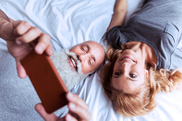 Älteres Paar liegt lächelnd im Bett und macht ein Selfie