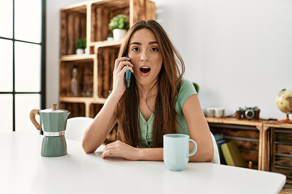 Eine Frau sitzt an einem Tisch und spricht erschrocken in ihr Smartphone